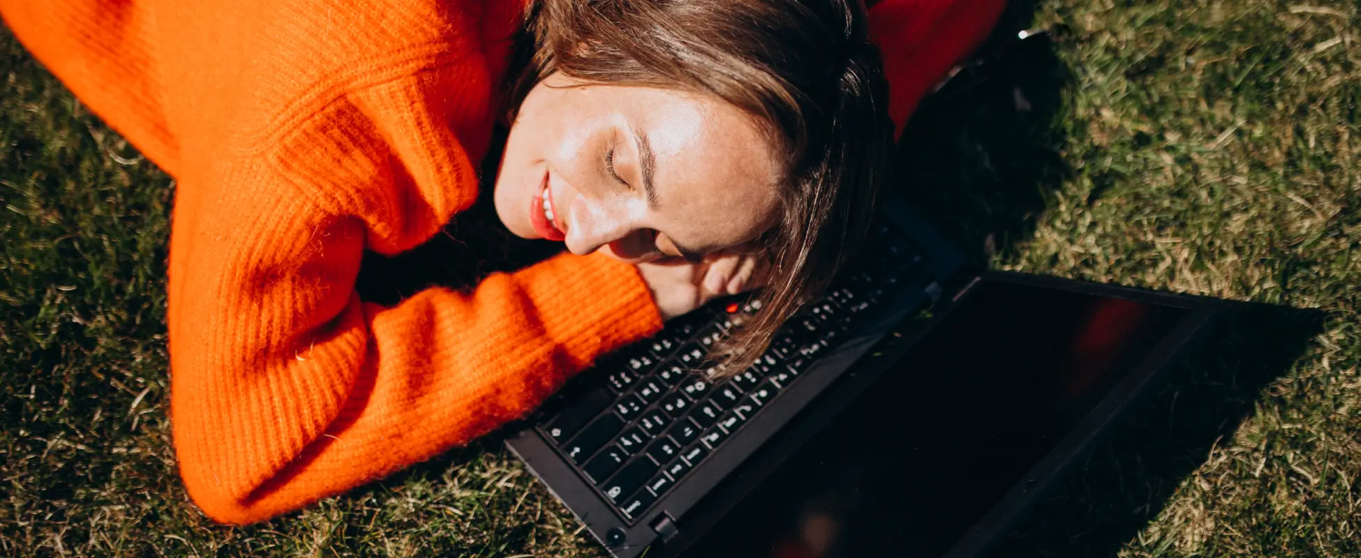 Woman lying on grass with her head resting on a laptop, taking a break from screens