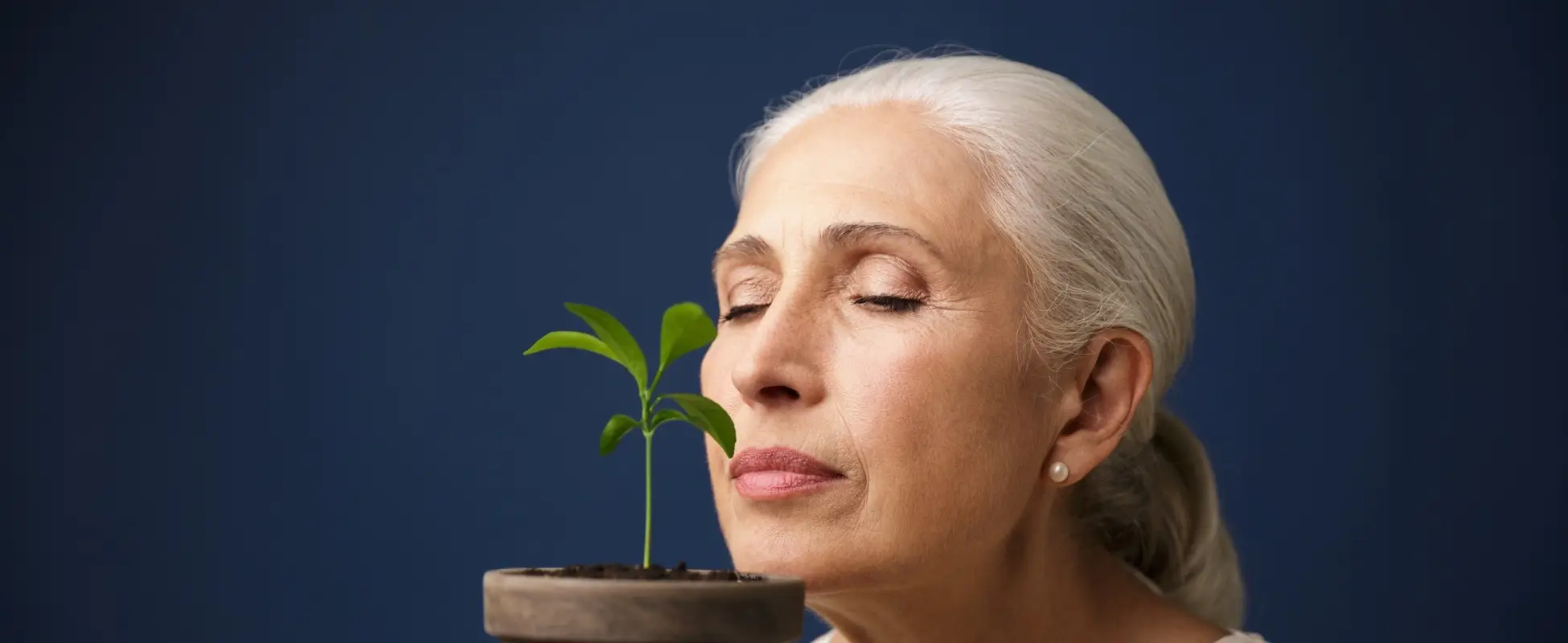 Regenerative Wellness vs Longevity vs Preventive Health Grey-haired woman with eyes closed smelling a green seedling in a clay pot, symbolising regenerative wellness and longevity